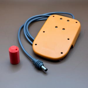 Op de afbeelding: Een close-up van een oranje voetpedaal met een zwart snoer en een rode stekker. Het pedaal heeft meerdere gaten en een getextureerd oppervlak.