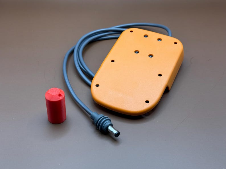 Op de afbeelding: Een close-up van een oranje voetpedaal met een zwart snoer en een rode stekker. Het pedaal heeft meerdere gaten en een getextureerd oppervlak.