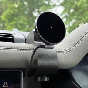 May include: A black wireless phone charger attached to a car's dashboard. The charger features a circular charging pad with a lightning bolt symbol. A black cable links the charger. The dashboard is grey.