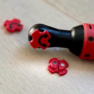 Puede incluir: Una herramienta de carraca eléctrica roja y negra con tres piezas de ajuste direccionales rojas. La herramienta está sobre una superficie clara con vetas de madera. Las piezas de ajuste tienen una flecha negra que indica la dirección.