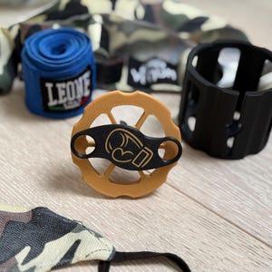 Puede incluir: Un surtido de equipamiento de boxeo, que incluye una venda azul para las manos con el texto "LEONE", una copa negra y un diseño de guante de boxeo marrón y negro. También se ve tela de camuflaje.