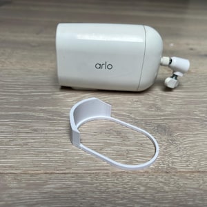 Könnte beinhalten: Weiße Arlo-Sicherheitskamera mit einer weißen Montagehalterung. Die Kamera hat ein weißes Kunststoffgehäuse mit dem Wort "arlo" auf der Vorderseite.