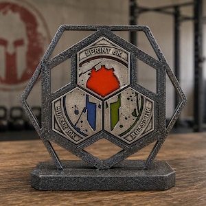 May include: A metallic gray award with a geometric design, featuring three inset panels with text and colorful graphics. The top panel reads "SPRINT 6K" above a red shape. The other panels read "SUPER 10K" and "BEAST 21K". The background shows a Spartan logo.