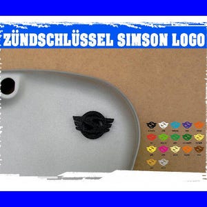 Op de afbeelding: Een close-up van een zilveren motorfietsonderdeel met een zwart embleem en de tekst "ZÜNDSCHLÜSSEL SIMSON LOGO" in het blauw. Een kleurenkaart met emblemen wordt hieronder weergegeven.