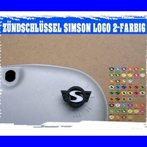 Op de afbeelding: Een close-up van een motorfietsonderdeel met de tekst "ZÜNDSCHLÜSSEL SIMSON LOGO 2-FARBIG". Het onderdeel is lichtgrijs met een zwart-wit logo. Een verscheidenheid aan gekleurde logo's wordt weergegeven.