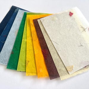 May include: A fan-shaped collection of rectangular handmade paper sheets in a spectrum of colours including dark blue, light blue, green, lime, yellow, orange, brown, and cream. Each sheet features a textured surface with embedded natural fibres.