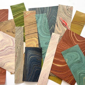 May include: A collection of rectangular paper sheets featuring various marbled patterns in shades of green, blue, brown, tan, pink, and cream. The paper surfaces display intricate, swirling, and wavy lines created by the traditional marbling technique.