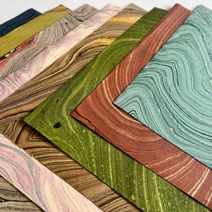 Könnte beinhalten: Eine Sammlung rechteckiger Papierbögen mit verschiedenen Marmormustern in den Farben Petrol, Terrakotta, Olivgrün, Gold, Lavendel und Dunkelblau, die in einer fächerartigen, überlappenden Anordnung mit wirbelnden Linien liegen.