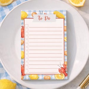 May include: A rectangular notepad with a blue and white checkered border, featuring illustrations of lemons, corn, potatoes, and crawfish. The notepad has a red lined section for a to-do list and the words "To Do" at the top.
