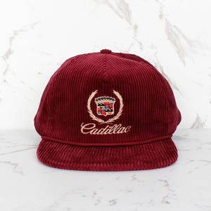 May include: A burgundy corduroy cap featuring the Cadillac emblem. The emblem includes a crest with a crown and the word "Cadillac" in gold script. The cap has a curved peak and a rounded crown. The cap is a deep red colour.