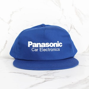 May include: A blue baseball cap with the white text "Panasonic Car Electronics" across the front. The cap has a curved brim and a button on top. The background is a white marble surface.