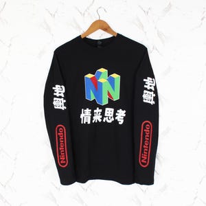 Könnte beinhalten: Ein schwarzes Langarmshirt mit einem bunten N64-Logo auf der Brust und weißem japanischen Text darunter. Beide Ärmel zeigen weiße japanische Schriftzeichen und ein rotes Nintendo-Logo. Das Shirt hängt an einem hölzernen Kleiderbügel.