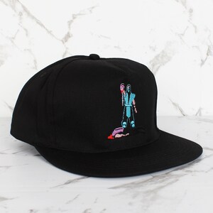 May include: Black baseball cap with an embroidered image of a video game character holding a weapon and standing over a fallen opponent.