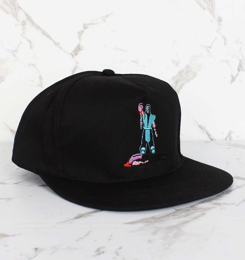 May include: Black baseball cap with an embroidered image of a video game character holding a weapon and standing over a fallen opponent.