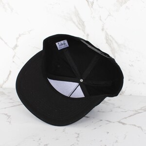 Op de afbeelding: Zwarte baseballpet met een zwarte klep en een zwarte kroon. De pet heeft een wit label aan de binnenkant met de tekst "FLY" en "SUPER FLY CLOTHING" in zwart.