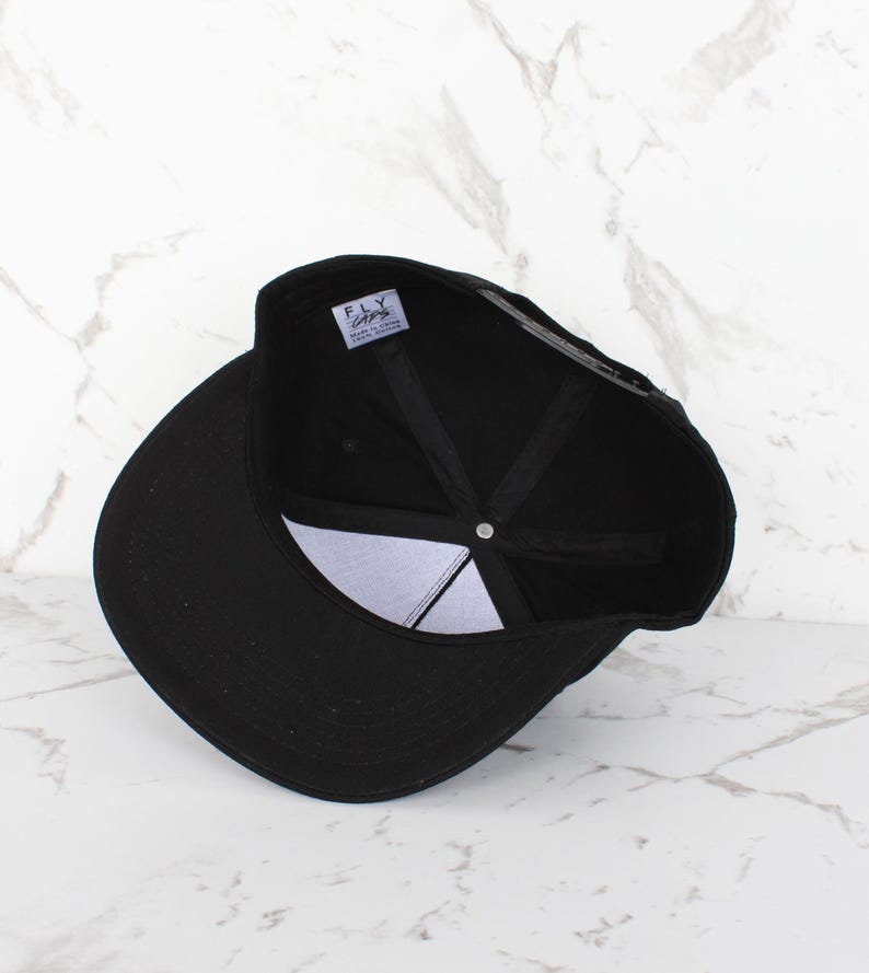 Op de afbeelding: Zwarte baseballpet met een zwarte klep en een zwarte kroon. De pet heeft een wit label aan de binnenkant met de tekst "FLY" en "SUPER FLY CLOTHING" in zwart.