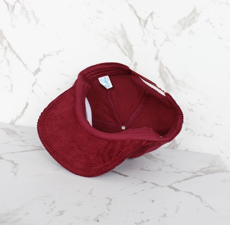 May include: A burgundy corduroy baseball cap. The cap features a curved peak and a structured crown. The interior of the cap is visible, showing a white tag and a small label. The cap is made of a ribbed corduroy fabric.