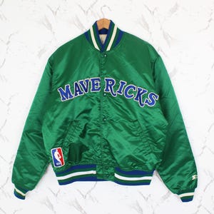 May include: Green satin jacket with the word "MAVERICKS" in blue and white lettering across the chest. The jacket has a blue, white, and green striped collar, cuffs, and hem. An NBA logo patch is on the left sleeve.