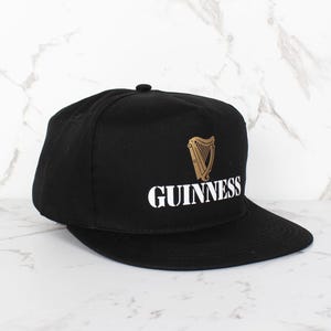 Op de afbeelding: Een zwarte en gouden baseballpet met het Guinness-logo en een harpdesign.