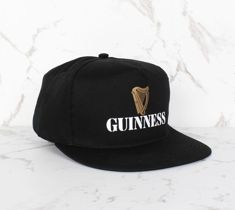 Op de afbeelding: Een zwarte en gouden baseballpet met het Guinness-logo en een harpdesign.