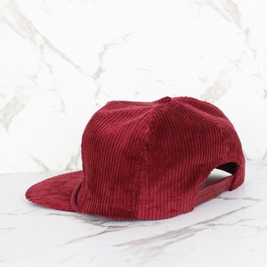 May include: A burgundy corduroy baseball cap with a flat peak. The cap features a structured crown and an adjustable strap at the back. The fabric has a ribbed texture.
