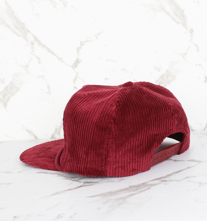 May include: A burgundy corduroy baseball cap with a flat peak. The cap features a structured crown and an adjustable strap at the back. The fabric has a ribbed texture.