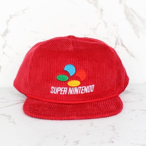 May include: Red corduroy baseball cap with the Super Nintendo logo embroidered on the front. The logo features a colorful design with the words "SUPER NINTENDO" in white. The cap has a curved brim.