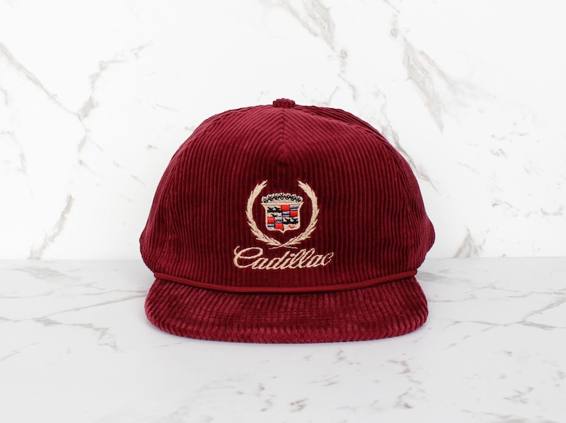 May include: A burgundy corduroy cap featuring the Cadillac emblem. The emblem includes a crest with a crown and the word "Cadillac" in gold script. The cap has a curved peak and a rounded crown. The cap is a deep red colour.