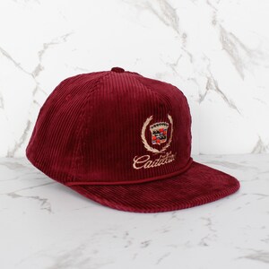 May include: A burgundy corduroy baseball cap with a flat peak. The cap features an embroidered Cadillac emblem in gold and red, with the brand name in cursive script below the emblem. The cap has a structured crown and a matching burgundy peak.