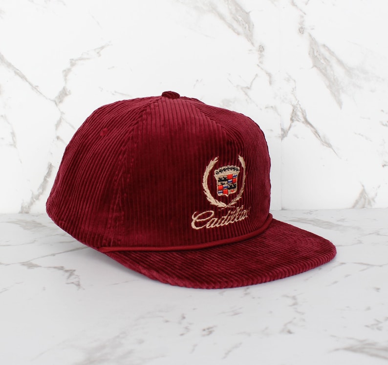 May include: A burgundy corduroy baseball cap with a flat peak. The cap features an embroidered Cadillac emblem in gold and red, with the brand name in cursive script below the emblem. The cap has a structured crown and a matching burgundy peak.