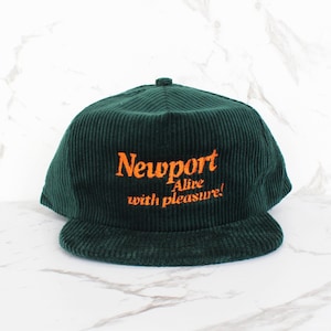 May include: A dark green corduroy baseball cap featuring the word "Newport" in orange script, and the phrase "Alive with pleasure!". The cap has a curved peak and a button. It sits on a white marble surface.