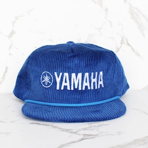 Puede incluir: Gorra de béisbol de pana azul con el logotipo blanco de Yamaha bordado en la parte delantera. La gorra tiene una visera curva y un detalle de cuerda azul. Estilo clásico.