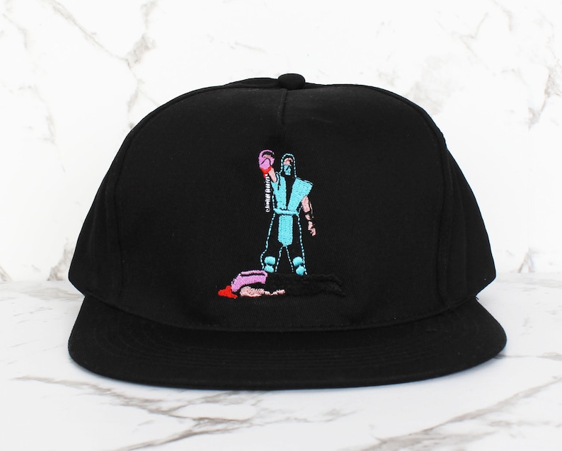 May include: Black and blue embroidered baseball cap with a video game character graphic. The character is wearing a blue and white outfit and is holding a pink fist.