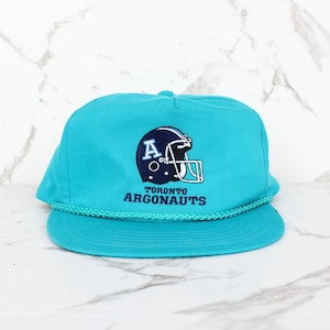 May include: A bright turquoise baseball cap featuring an embroidered dark blue football helmet with a white letter A on the side. Below the helmet, the text TORONTO ARGONAUTS is embroidered in dark blue. A matching turquoise braided cord trim sits above the brim.
