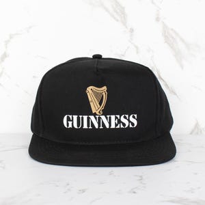 Op de afbeelding: Zwarte baseballpet met een wit "GUINNESS" logo en een gouden harp afbeelding.