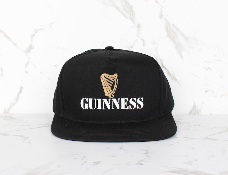 Op de afbeelding: Zwarte baseballpet met een wit "GUINNESS" logo en een gouden harp afbeelding.