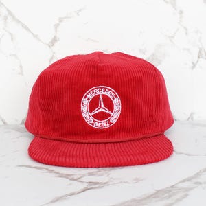 May include: Red corduroy baseball cap with a white embroidered Mercedes-Benz logo.