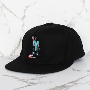 May include: Black baseball cap with an embroidered image of a video game character holding a weapon. The character is wearing a blue and white outfit and is standing over a fallen opponent.