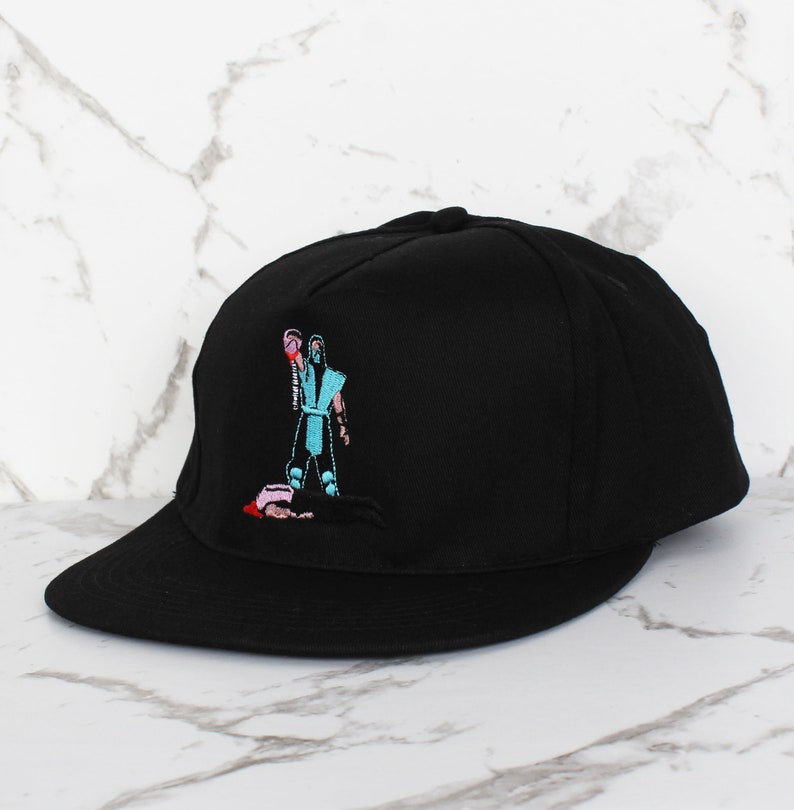 May include: Black baseball cap with an embroidered image of a video game character holding a weapon. The character is wearing a blue and white outfit and is standing over a fallen opponent.