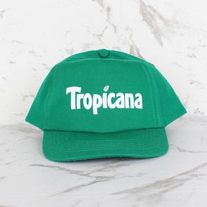May include: A green baseball cap featuring the white word "Tropicana". The cap has a curved peak and a button on top. The hat is made of a fabric material. The cap is a vibrant green colour.