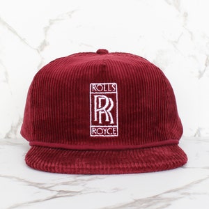 Puede incluir: Una gorra de béisbol de pana roja con un logotipo de Rolls Royce bordado en blanco.