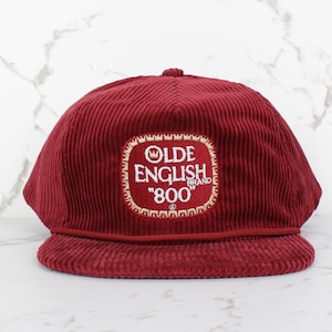 Puede incluir: Gorra de béisbol de pana burdeos con visera curva. La gorra presenta un parche bordado con las palabras "OLDE ENGLISH BRAND 800" en blanco y dorado. La gorra tiene una corona estructurada y una banda burdeos a juego.