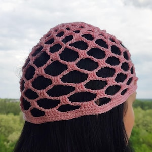 May include: A light pink crochet beanie featuring an openwork net pattern with large, irregular diamond-shaped holes and a solid, tightly stitched band around the bottom edge that sits securely against the head.