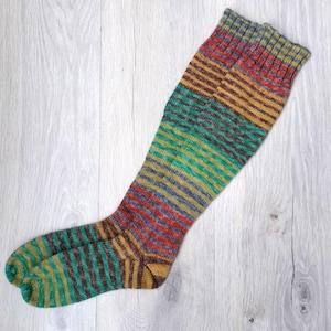 May include: A pair of knee-high socks with horizontal stripes in shades of red, brown, yellow, green, and blue. The socks are made of a knitted material and are displayed on a light wood surface.
