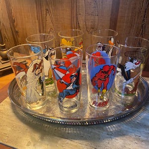 May include: A set of eight clear glass tumblers with cartoon character designs, including The Flash, Sylvester the Cat, and The Penguin. The glasses are arranged on a silver serving tray.