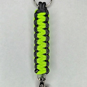Puede incluir: Un llavero de paracord negro y verde lima con un anillo de llave plateado y un clip plateado.