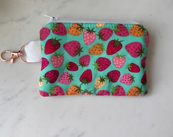 Peut inclure: Un petit porte-monnaie rectangulaire avec un fond turquoise et un motif de fraises rouges, roses et oranges. Il possède une sangle blanche avec un fermoir or rose et une fermeture éclair.