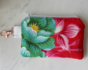 Peut inclure: Un petit porte-monnaie rectangulaire à imprimé floral avec fermeture éclair. Le sac à main présente un design vibrant avec des fleurs vertes, bleues et roses sur fond rouge. Il comprend une sangle blanche et un fermoir or rose.