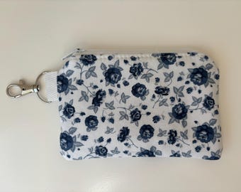 Peut inclure: Un porte-monnaie rectangulaire en tissu à fond blanc orné d'un motif répétitif de roses et de feuilles bleu foncé. La pochette est dotée d'une fermeture éclair blanche sur le bord supérieur et d'un mousqueton en métal argenté sur une patte blanche.