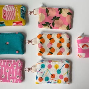 May include: A collection of small, zippered pouches with various colorful patterns. Designs include milk cartons, peaches, oranges, and spoons. Each pouch has a metal clasp and a fabric loop.